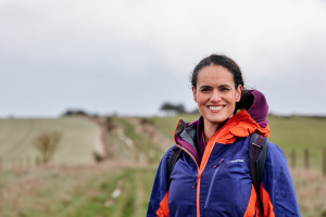 Mary-Ann wears a blue and orange raincoat and smiles at the camera. behind her there's a path that tracks to the horizon. the weather looks wet